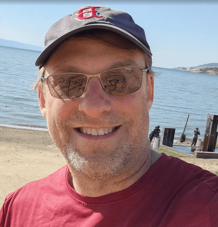 Smiling man wearing a baseball cap and sunglasses at a sunny beach.