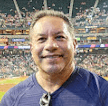 Smiling man in a blue shirt posing for a selfie at a crowded baseball stadium.